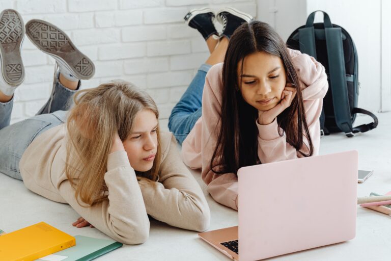 Two girls lying on their stomachs are looking intently at a computer laptop