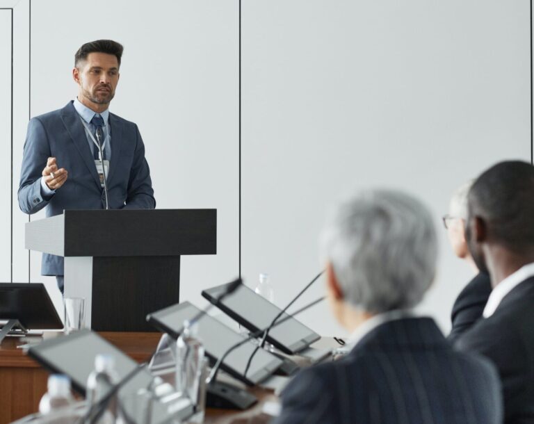 Man in suit at a podium talking to a panel of individuals sitting at a table