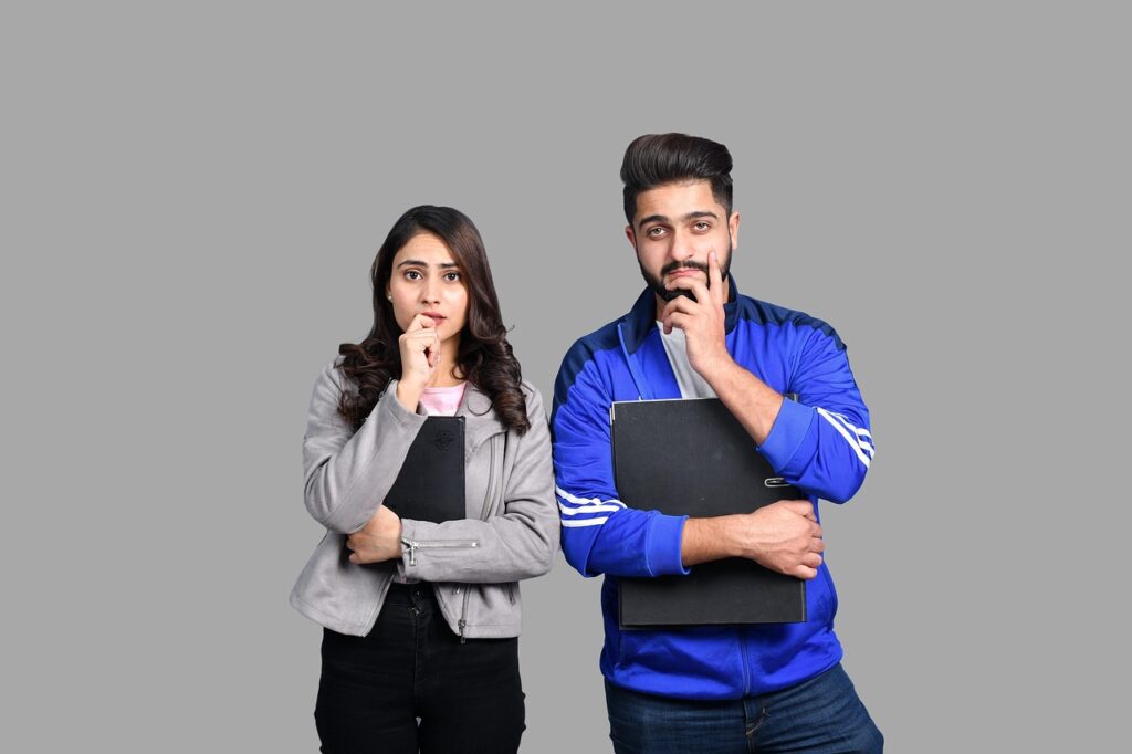 A young woman and man holding notebooks and facing the camera with one of their hands under their chins
