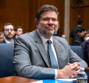 A smiling man dressed in a suit with hands folded and sitting at a desk with people behind him.