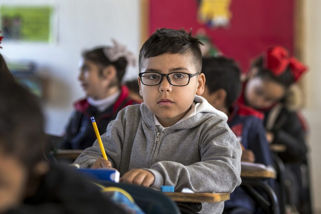 Hispanic boy wearing large glasses and holding a pencil while he sits at a desk in a classroom