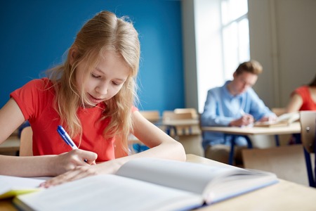 student girl with opened book completing a test on paper and pen